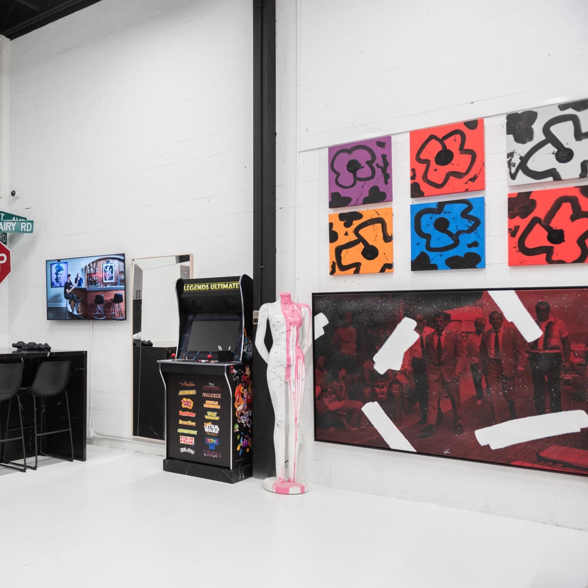 A modern gallery space with a bar area featuring stools, a stop sign, an arcade machine, and colorful artwork on the walls.