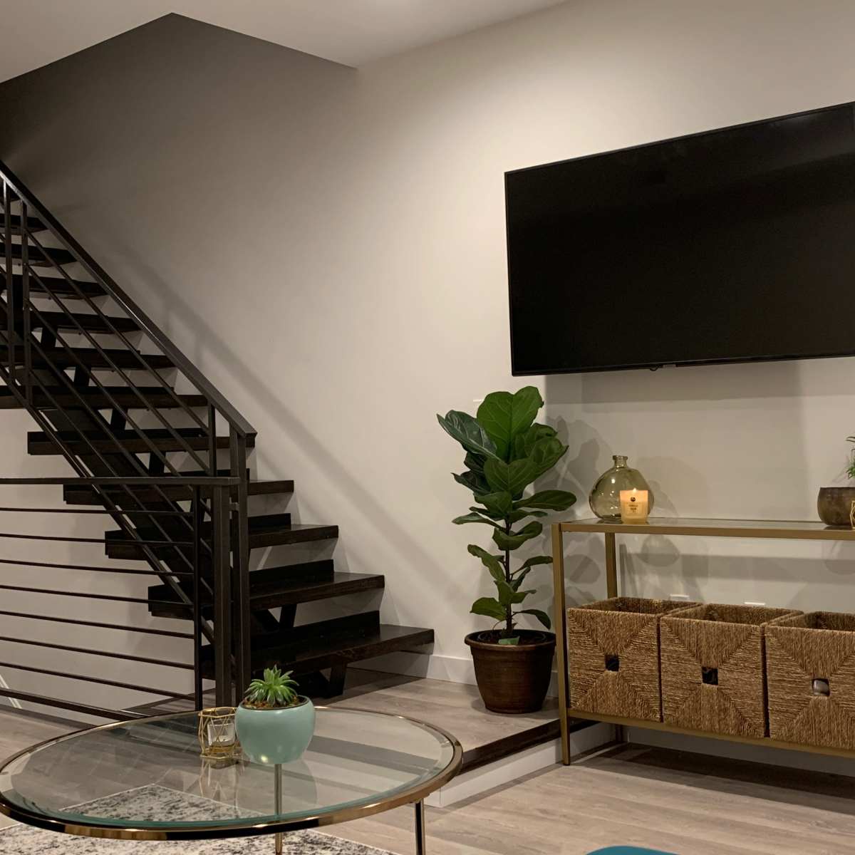 A modern living space features a staircase, a wall-mounted television, and a decorative shelf with storage baskets and plants.