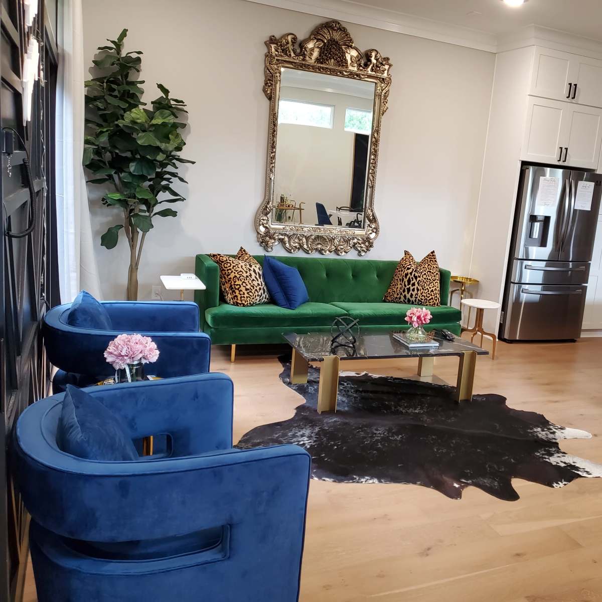 A modern living area featuring a green velvet sofa with patterned pillows, two blue chairs, a glass coffee table, and a kitchen area in the background.