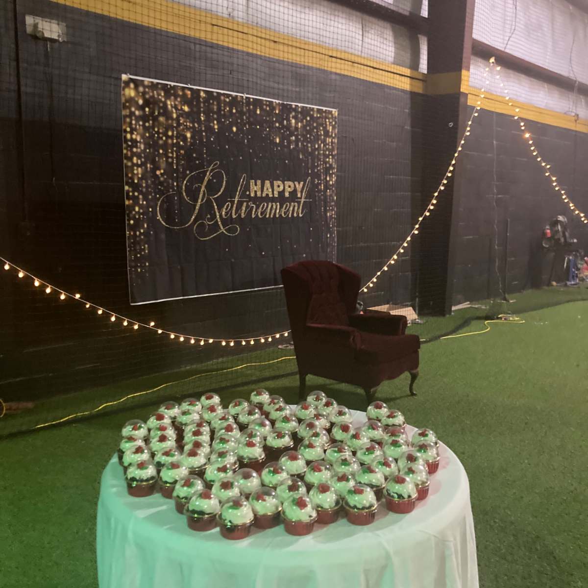A table covered with a white cloth displays numerous cupcakes in front of a "Happy Retirement" banner illuminated by string lights in a decorated venue.