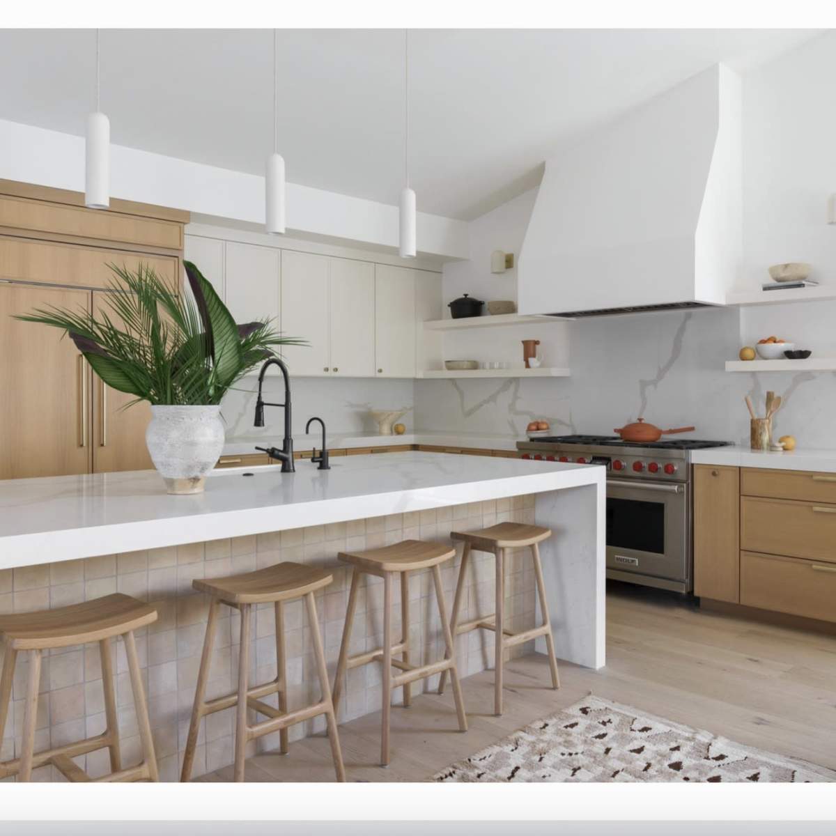 A modern kitchen features a white countertop island with bar stools, light wood cabinets, and an array of cooking appliances against a marble backsplash.