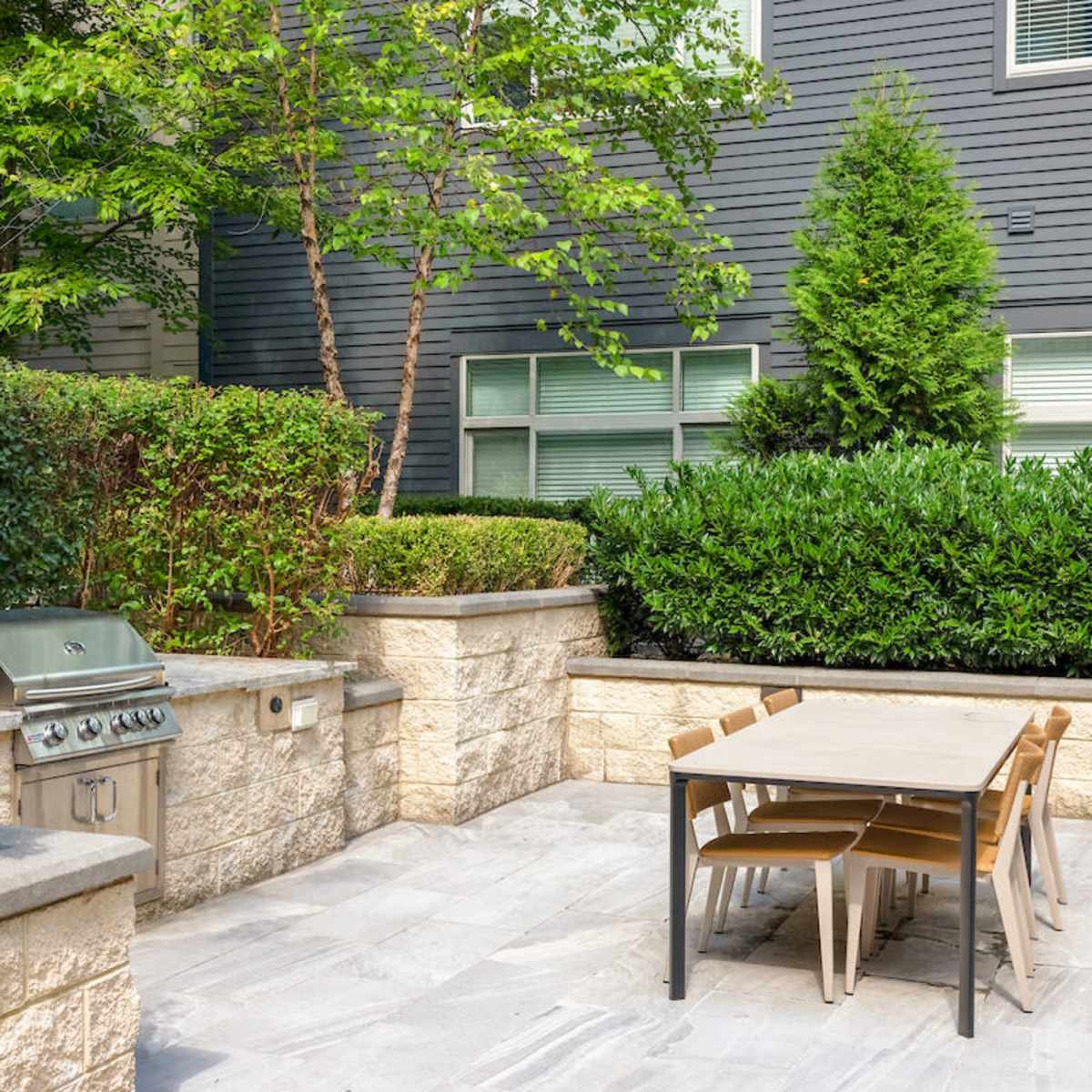 An outdoor patio area featuring a grill and a table surrounded by landscaped greenery and stone walls.