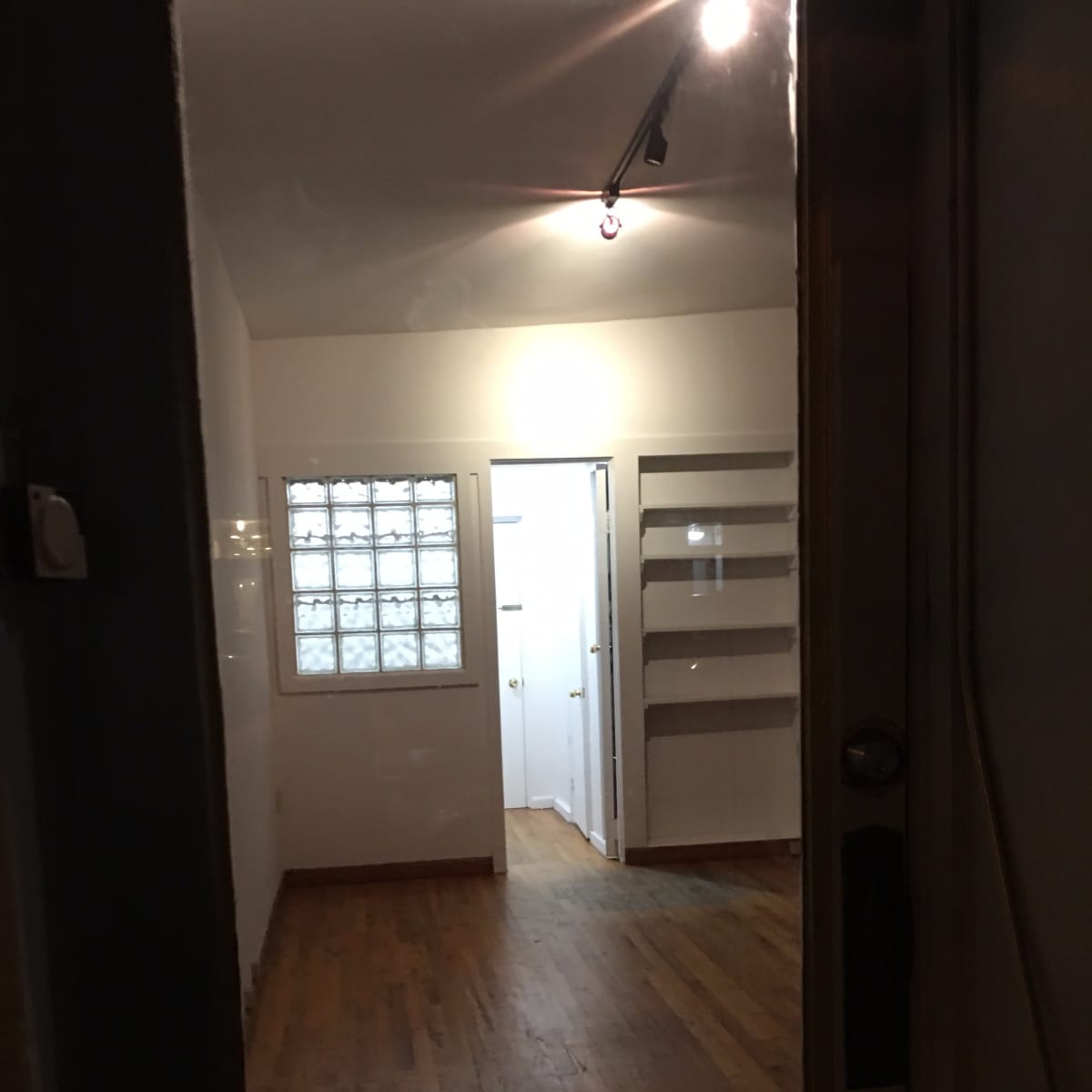 A wooden-floored room viewed through a glass door, with a hallway leading to another area and a light fixture overhead.