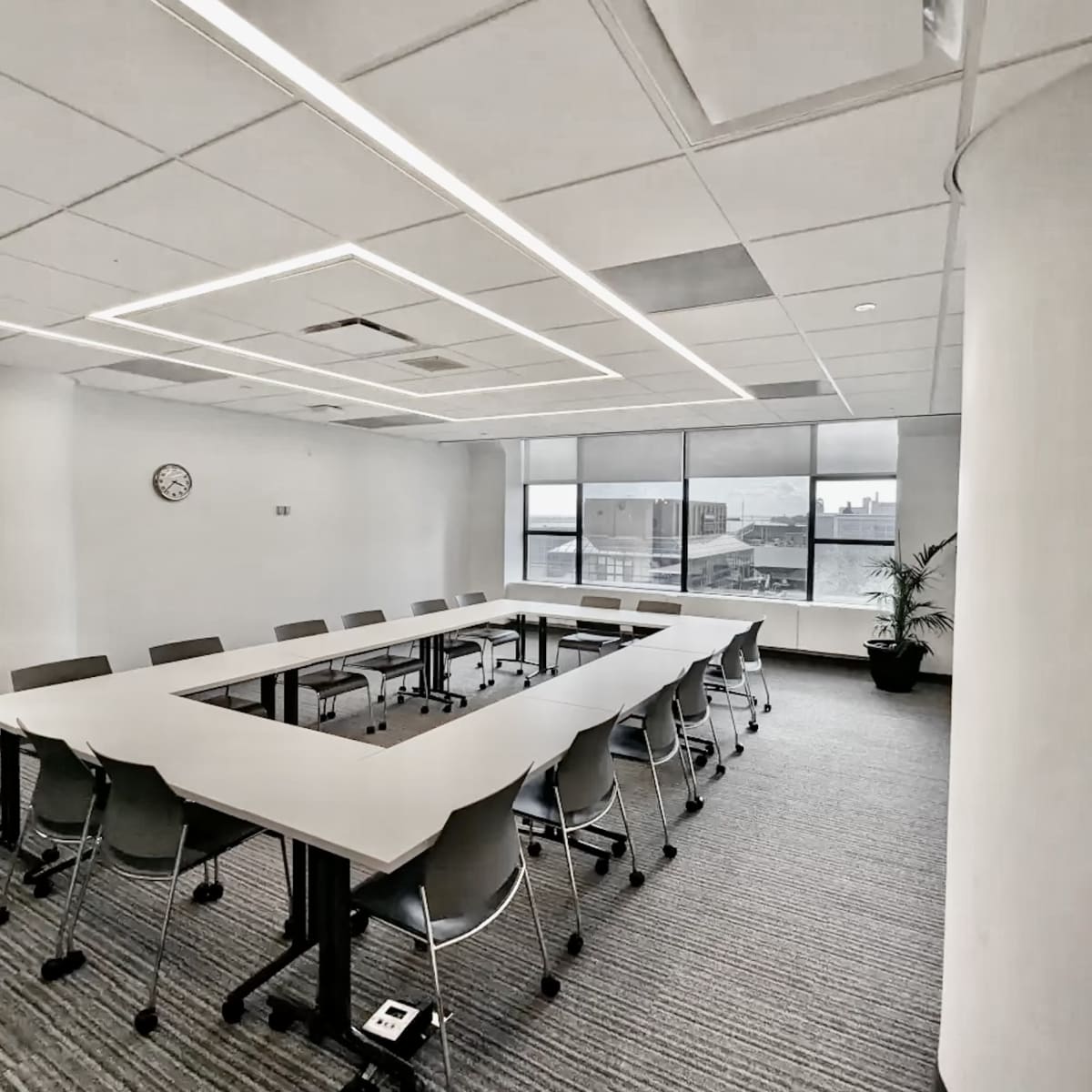 A modern conference room features a large rectangular table surrounded by chairs, with large windows providing natural light.