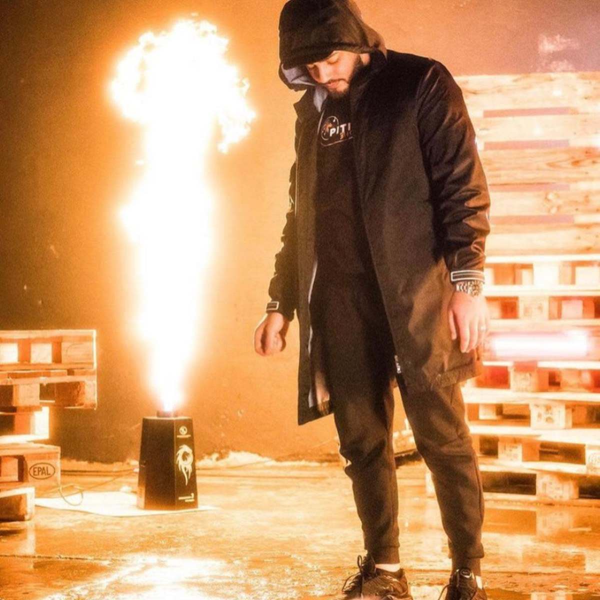 A man in a black hooded jacket and pants stands on a reflective surface in front of a flame shooting up from a speaker.