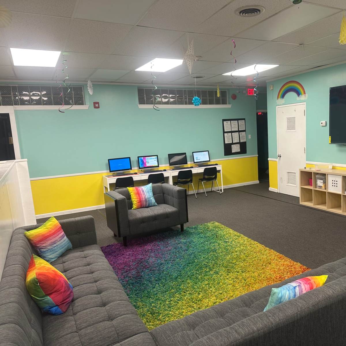 A brightly colored lounge area with a gray couch and chairs, a rainbow rug, computers along the wall, and a large screen displaying a video.