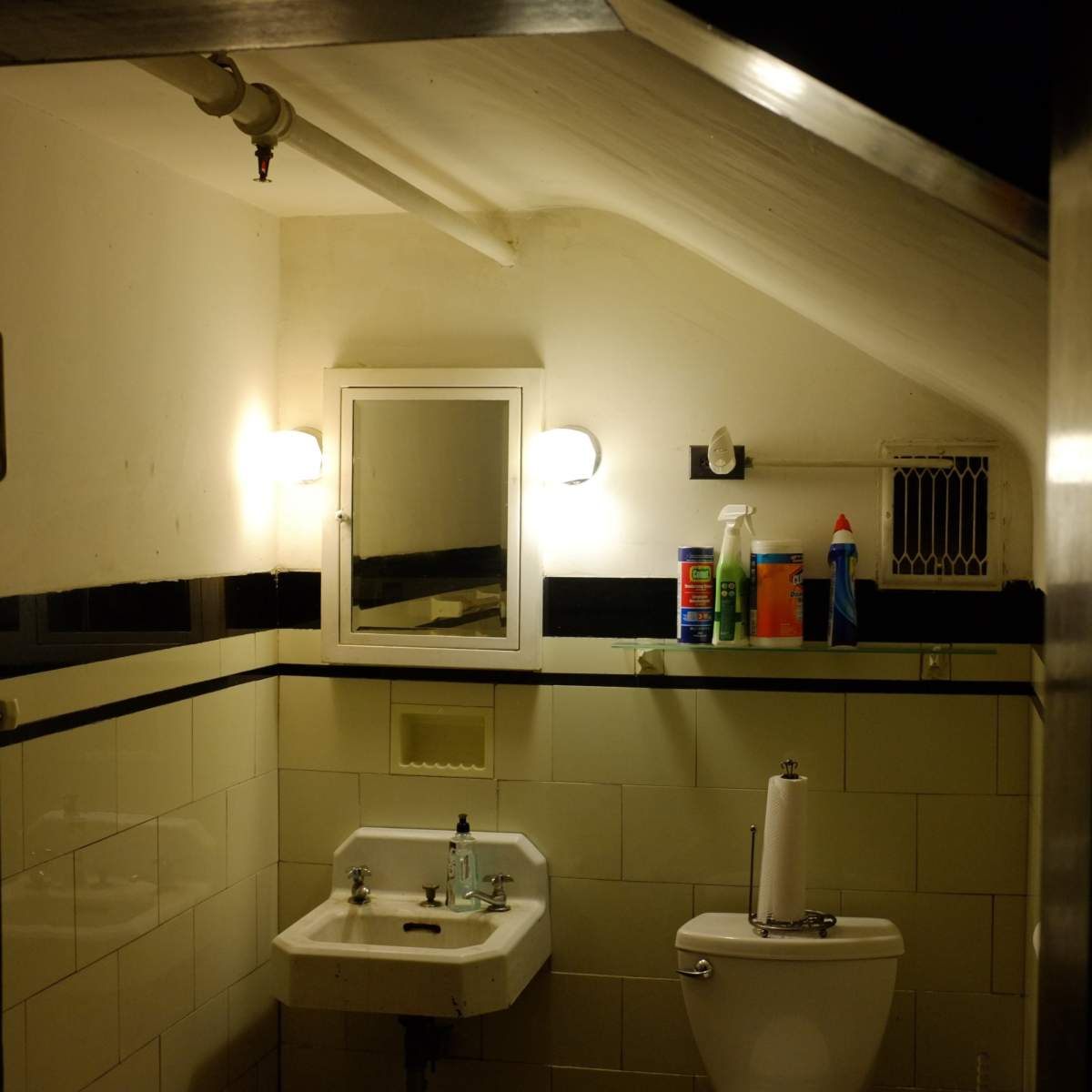 A small, sloped bathroom with a sink, toilet, and cleaning supplies on a shelf.