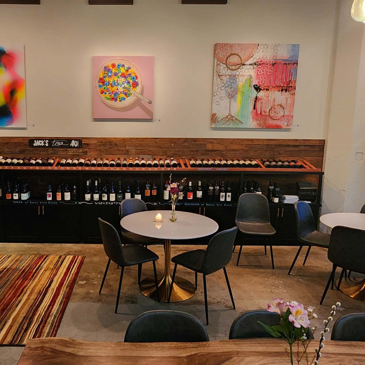 A modern wine bar interior with tables, artwork on the walls, and a display of wine bottles in the background.