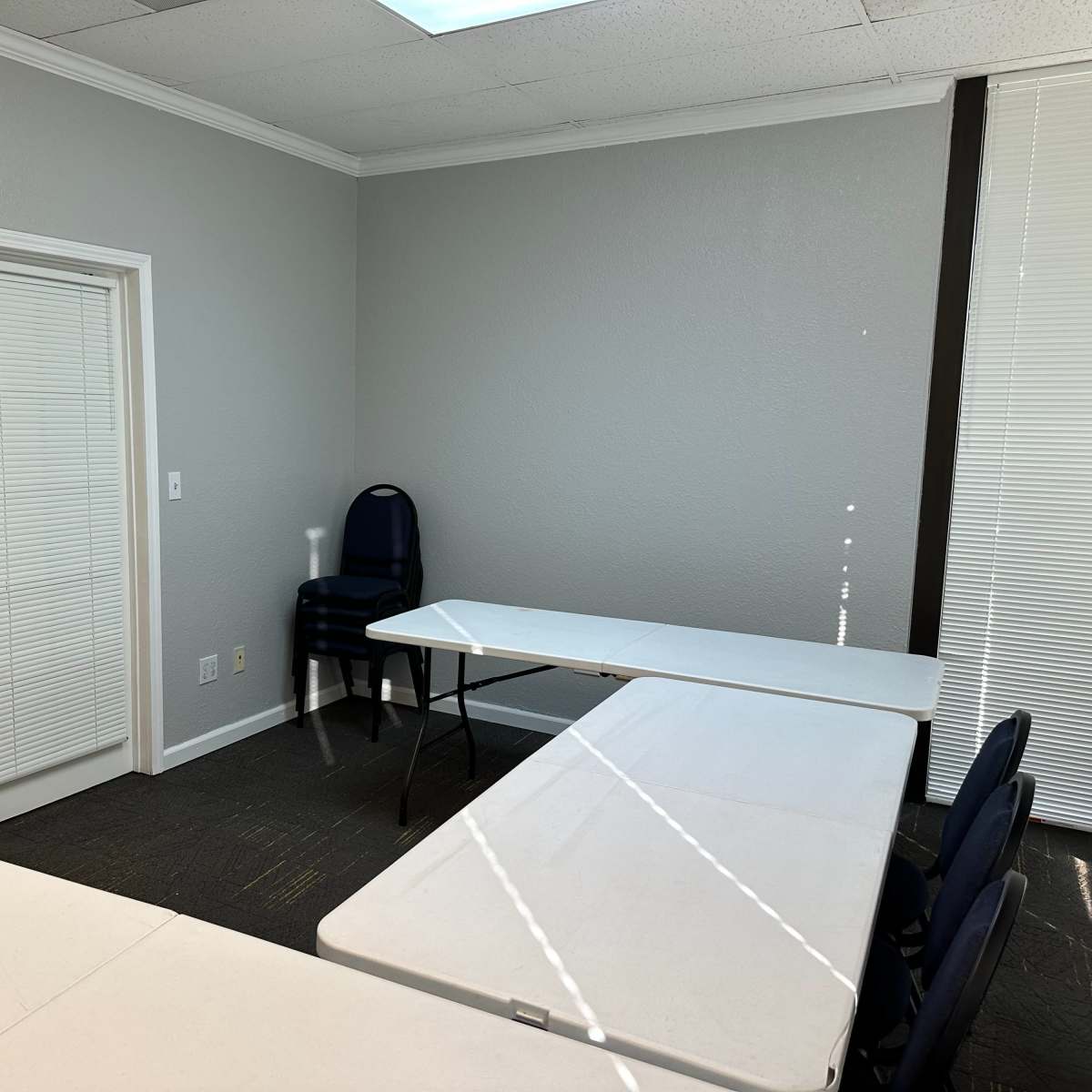 A small meeting room with two white tables arranged in an L-shape, a blue chair stacked against the wall, and a door leading to another room.
