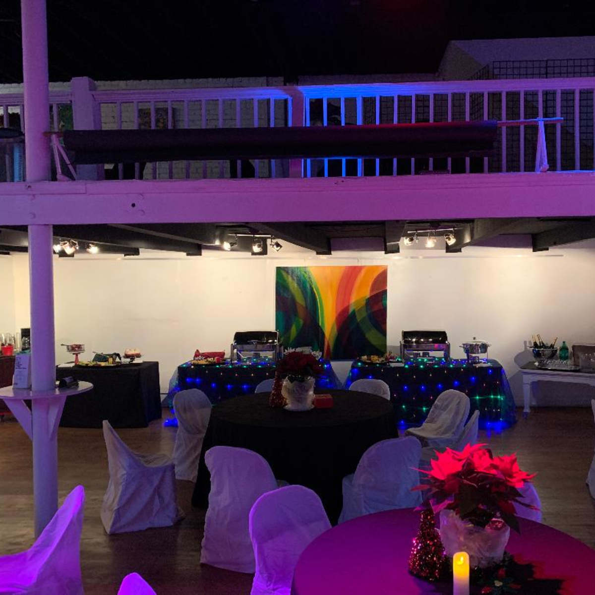 A banquet room is set up with tables adorned in black and purple tablecloths, featuring a colorful abstract painting on the wall and a buffet table filled with food.
