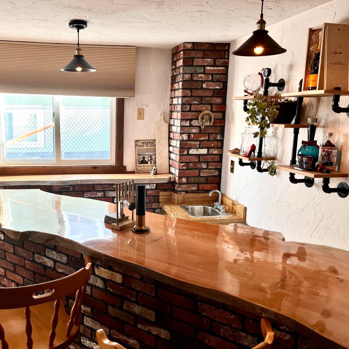 A small bar area with a curved wooden countertop, brick walls, and two wooden stools, illuminated by hanging light fixtures.