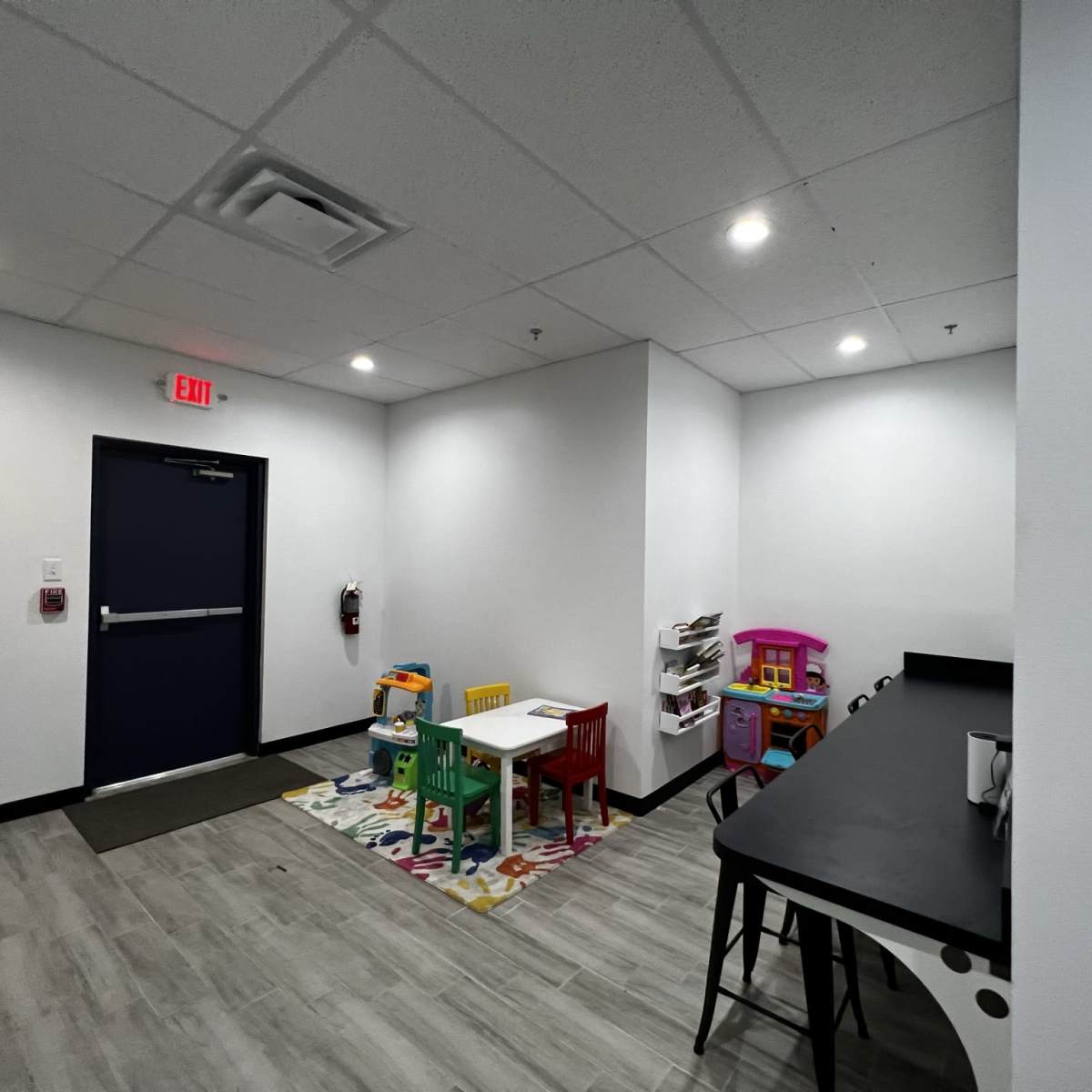 A brightly lit indoor play area featuring a small white table with colorful chairs, a toy kitchen set, and a playhouse on a patterned rug.