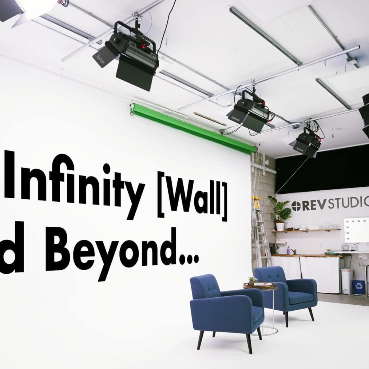 A brightly lit studio setup featuring a large white wall with the phrase "To Infinity [Wall] and Beyond..." and two blue chairs on a gray floor.