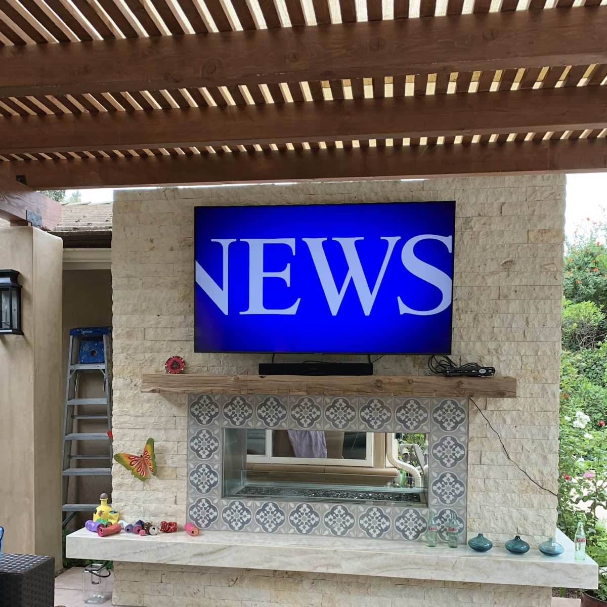 A television displaying the word "NEWS" is mounted on a stone wall above a decorative shelf in a patio area.