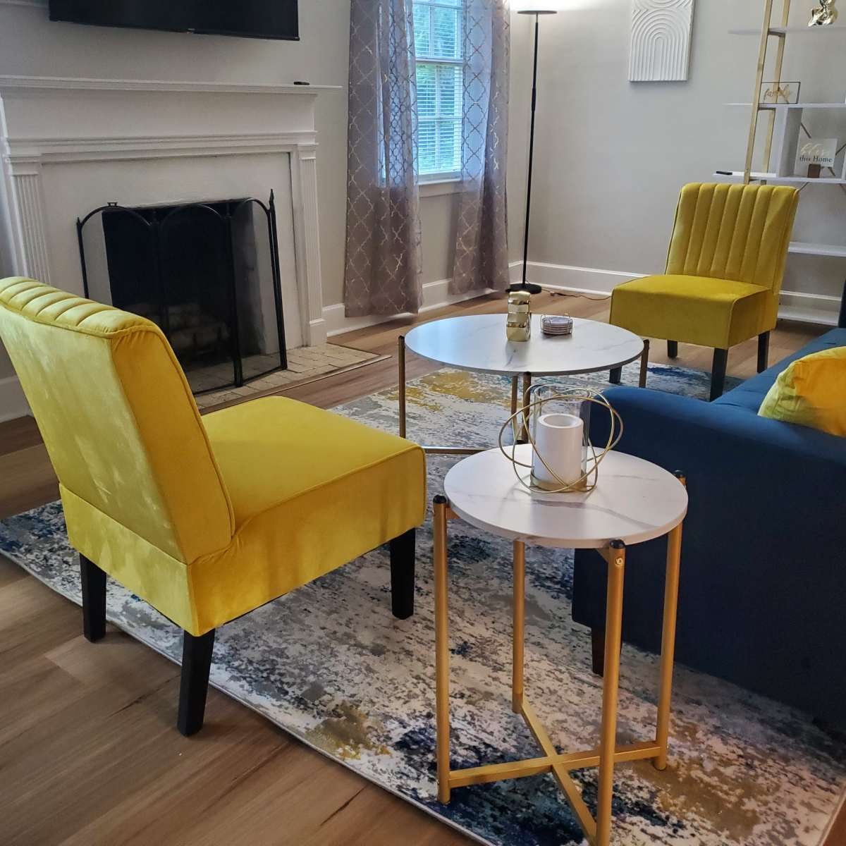 A modern living room with two yellow chairs and a blue sofa arranged around a small round table on a decorative rug, with a fireplace and a floor lamp in the background.