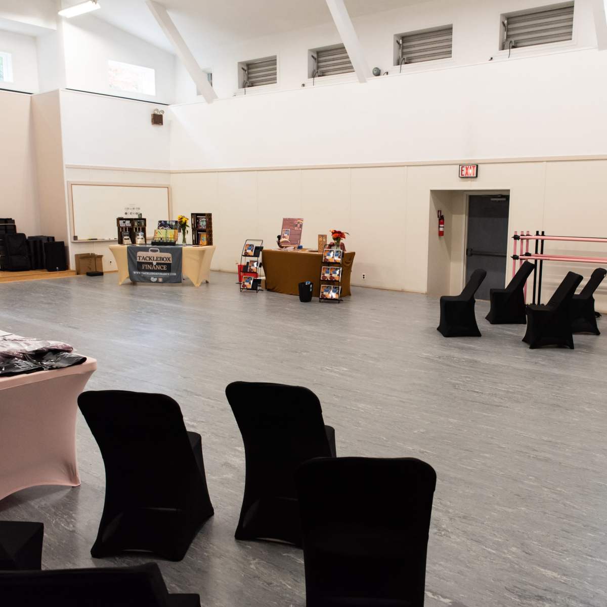 An empty room with a light gray floor, black chairs arranged in a circle, and various display tables set up along the walls.