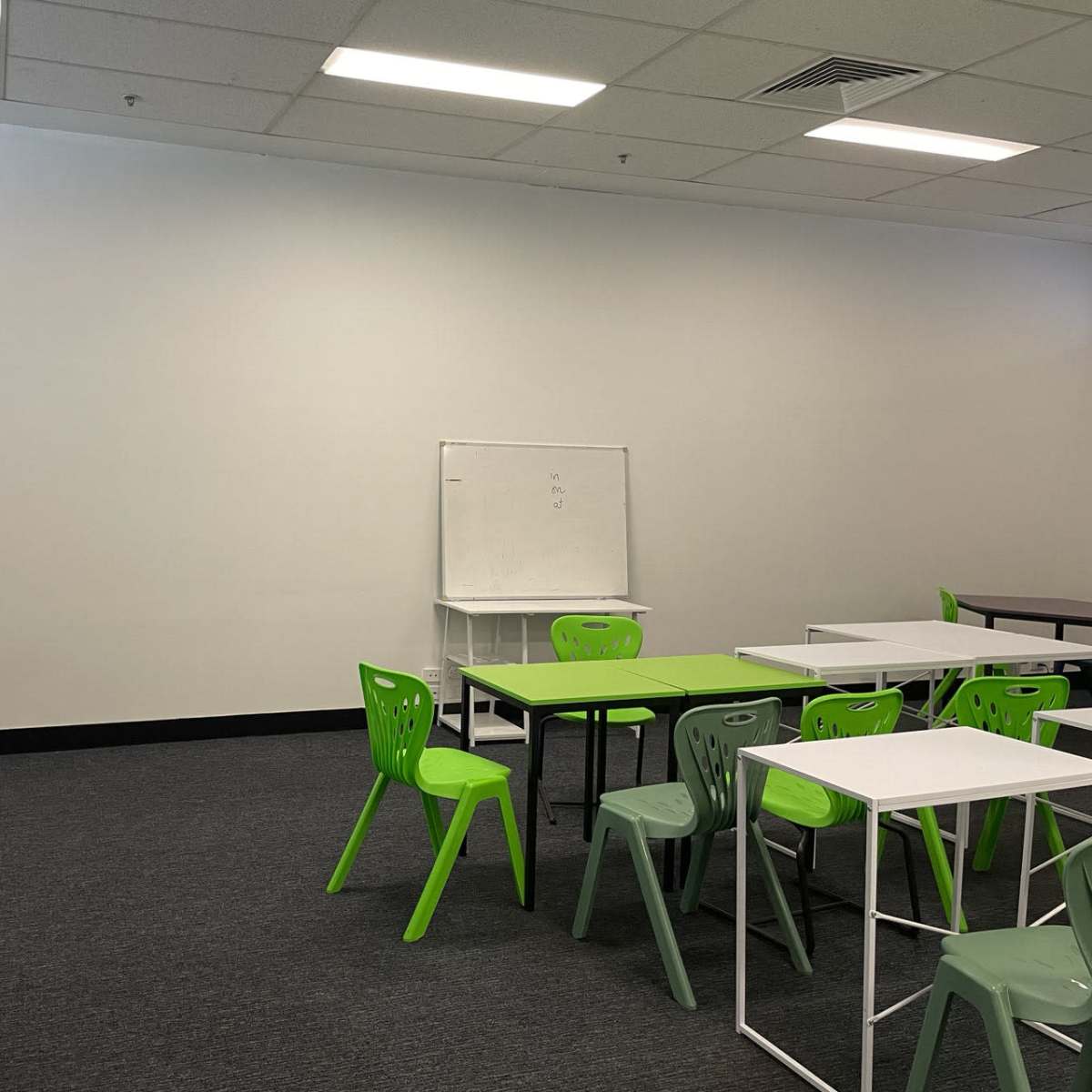 An empty classroom features green chairs and tables arranged against a plain wall with a whiteboard.
