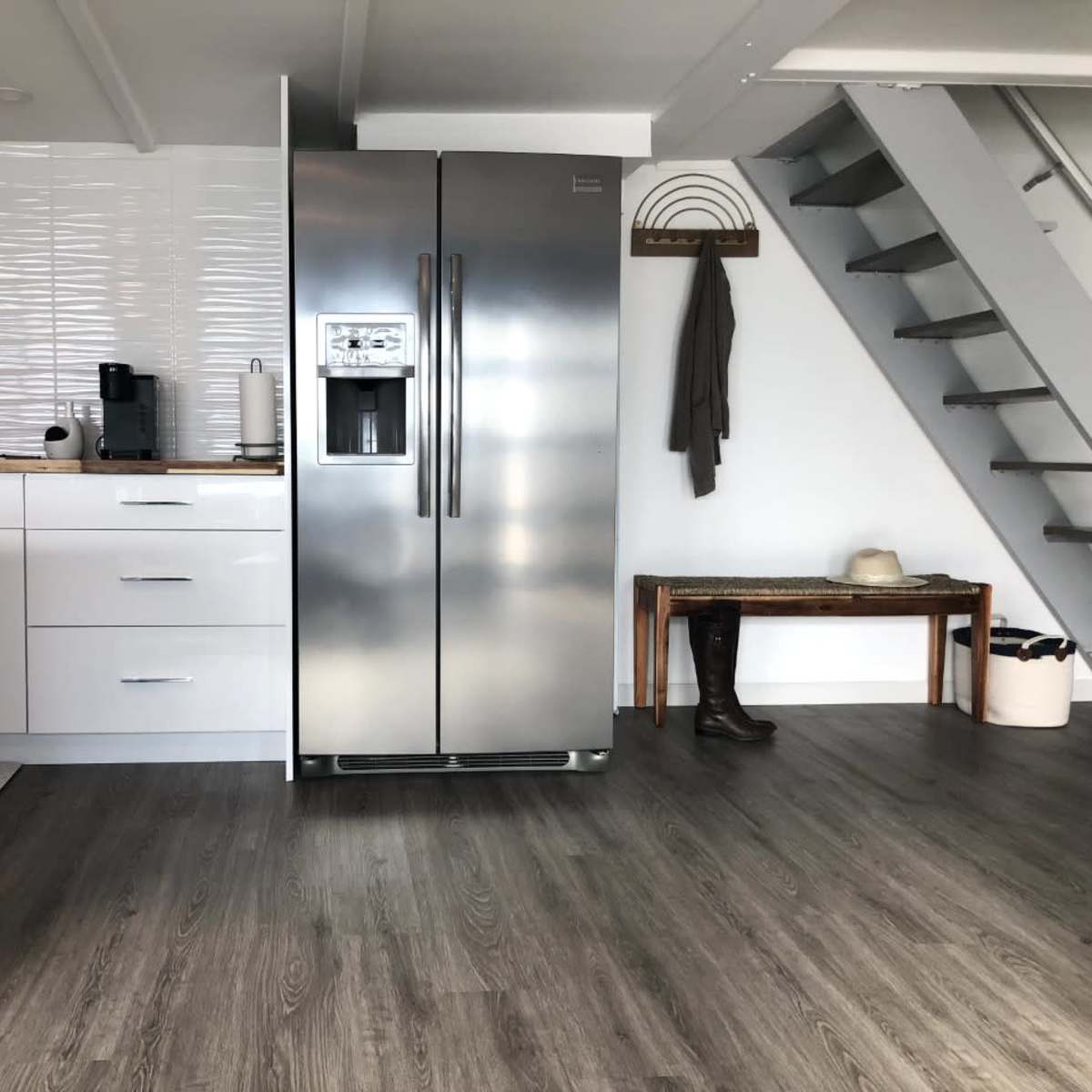 A modern kitchen area with a stainless steel refrigerator, wooden counter, and a staircase in a well-lit room.
