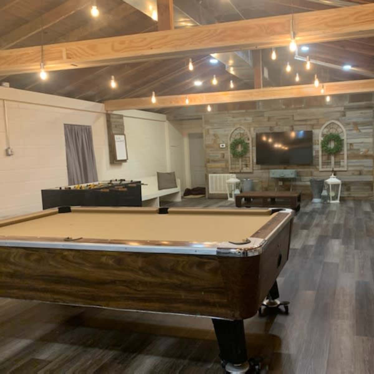 The room features a pool table, a foosball table, and a large television mounted on a wooden accent wall, with recessed lighting and wooden beams overhead.