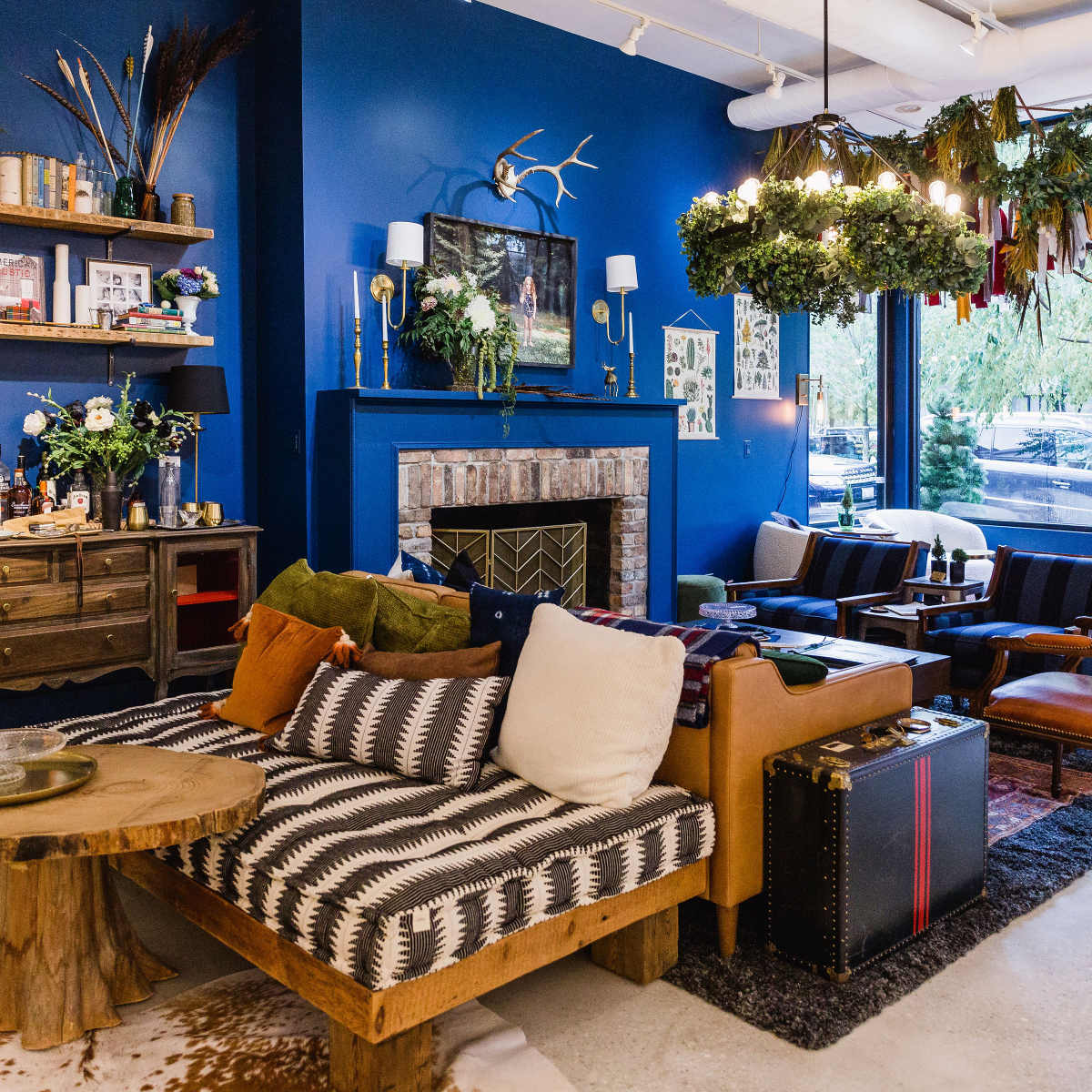 A stylish living room featuring a blue accent wall, a mix of wooden furniture, a fireplace, and greenery suspended from the ceiling.