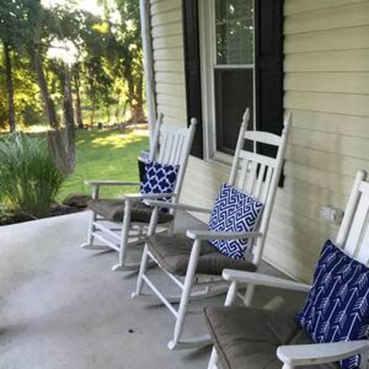 A porch with three white rocking chairs, each adorned with blue patterned cushions, overlooking a green yard.