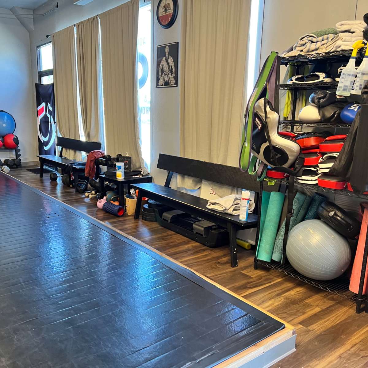 A fitness studio with a black mat area on the floor, benches along the side, and shelves stocked with various training equipment and exercise gear.
