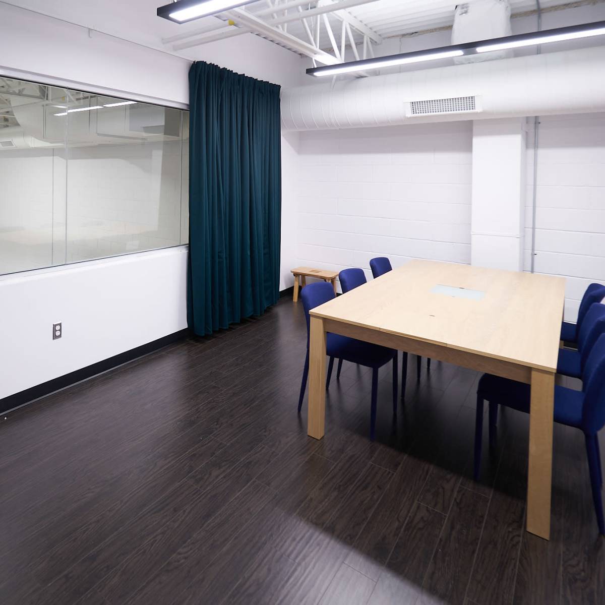 A modern meeting room with a large wooden table and blue chairs, featuring a glass wall that overlooks a separate space.