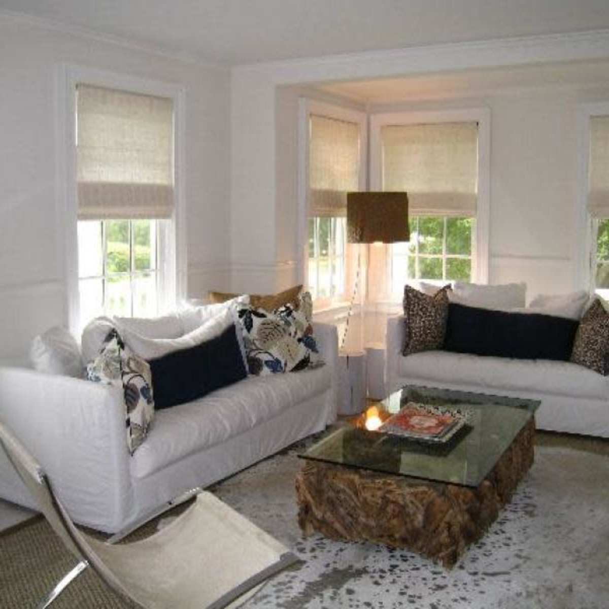 A light-filled living room features two white sofas flanking a glass coffee table on a patterned rug, with large windows and neutral window shades.