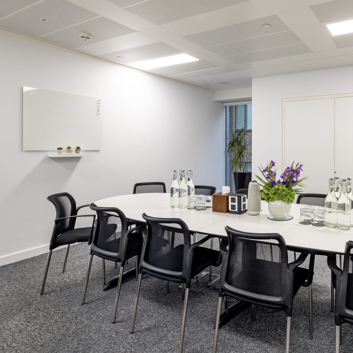 A conference room features a white oval table surrounded by black chairs, with glass water bottles and a flower arrangement in the center.