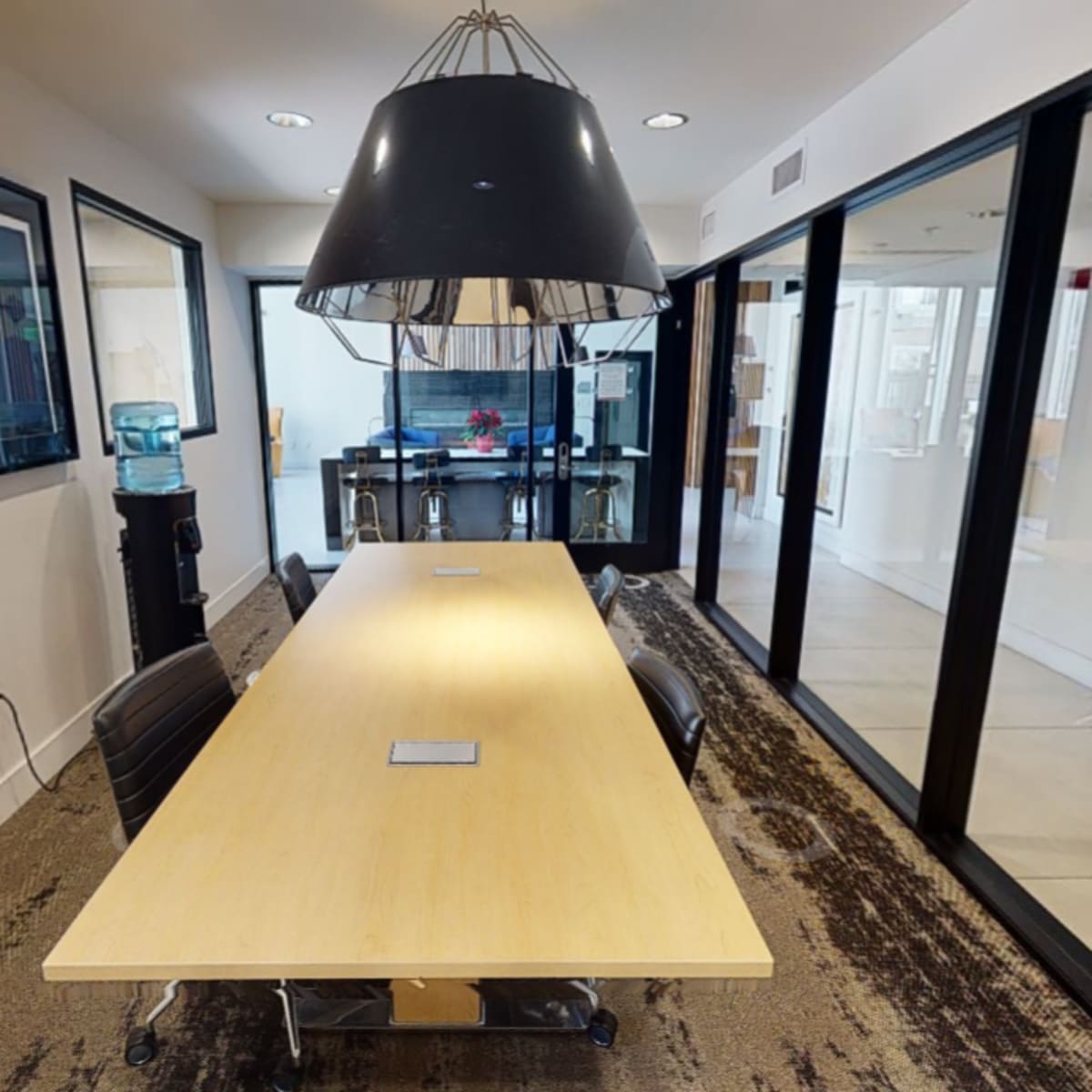 A modern conference room with a long wooden table, black chairs, and a large pendant light overhead, flanked by glass walls.