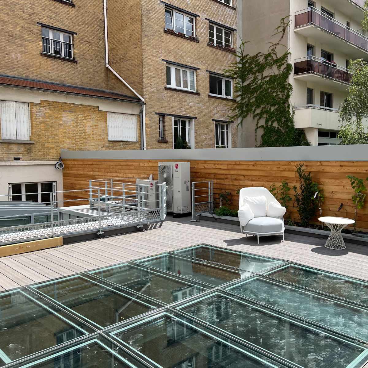 A rooftop terrace with two white armchairs and a small table, surrounded by wooden fencing and green plants.