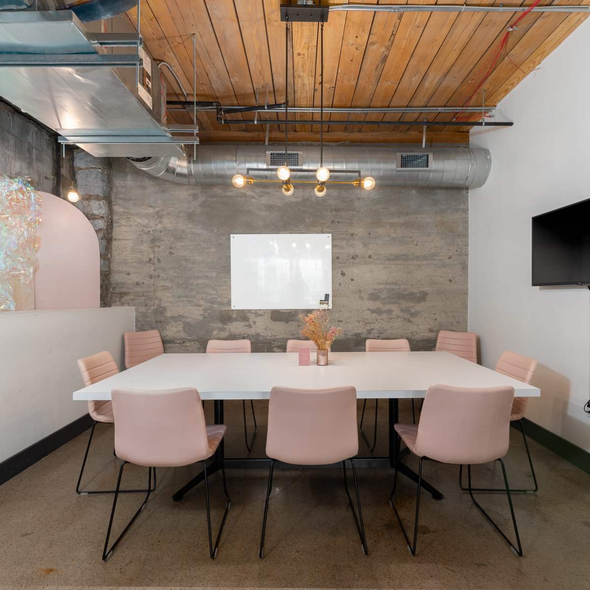 A modern conference room features a long white table surrounded by pink chairs, with exposed wooden beams and a large screen on the wall.