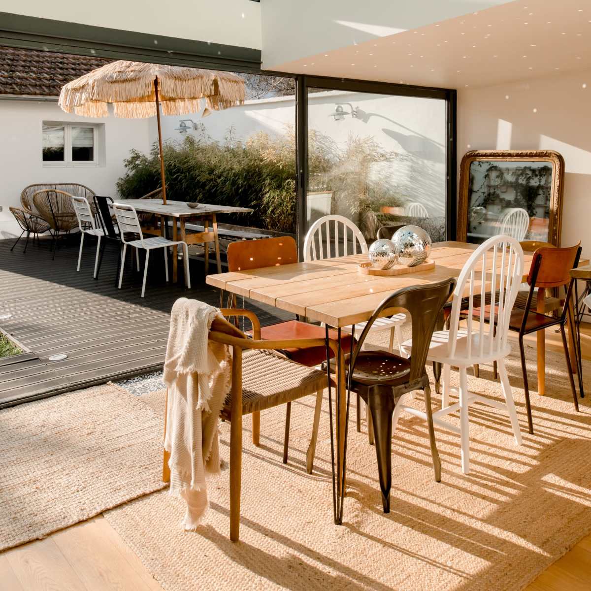 A bright, modern dining area with large windows, a woven rug, and a mix of wooden and metal chairs surrounding a wooden table.