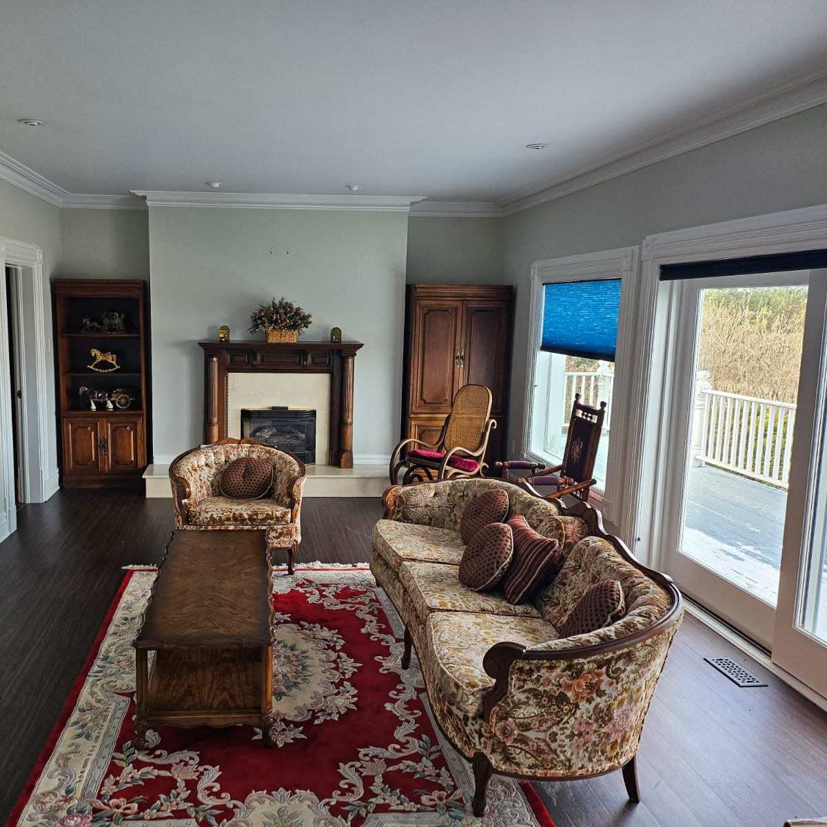 A living room with floral-patterned couches, a wooden coffee table, a fireplace flanked by wooden cabinets, and large windows with blue blinds.