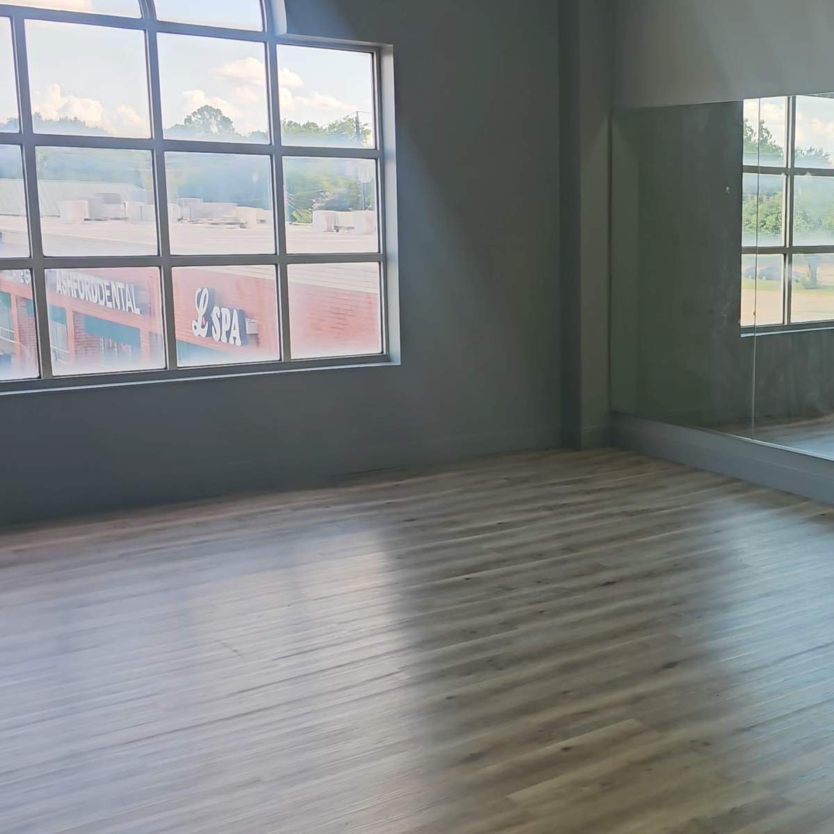 A spacious, well-lit room with wooden flooring, large windows, and a mirror covering one wall, along with a row of teal chairs positioned against another wall.