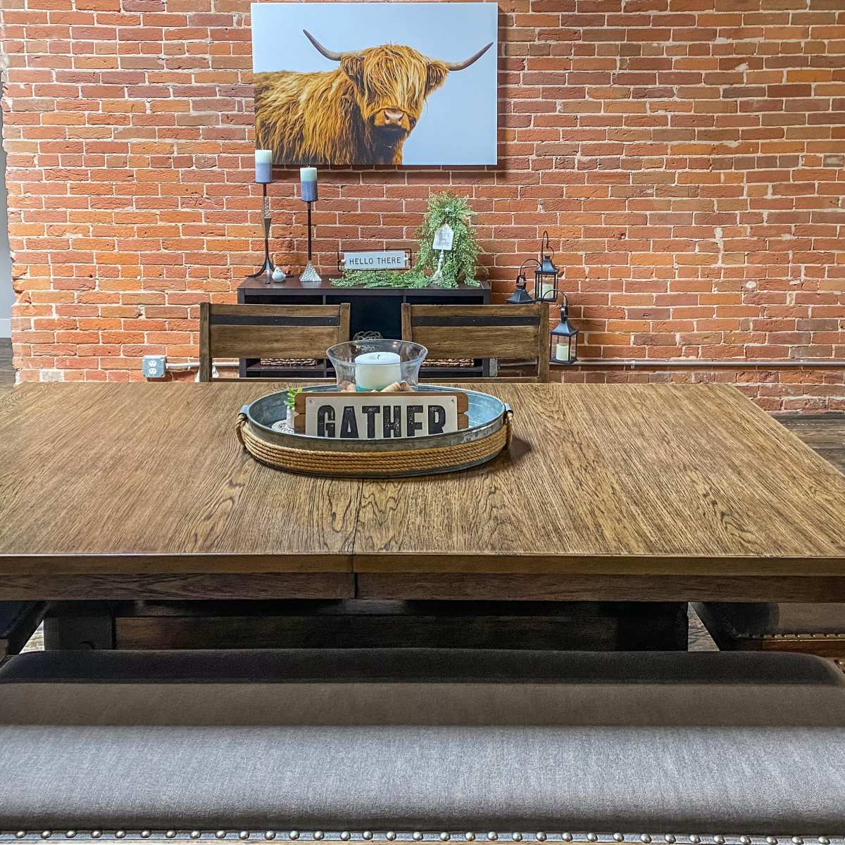 A wooden dining table with a decorative tray sits in front of a brick wall, accompanied by wooden chairs and a framed picture of a Highland cow.
