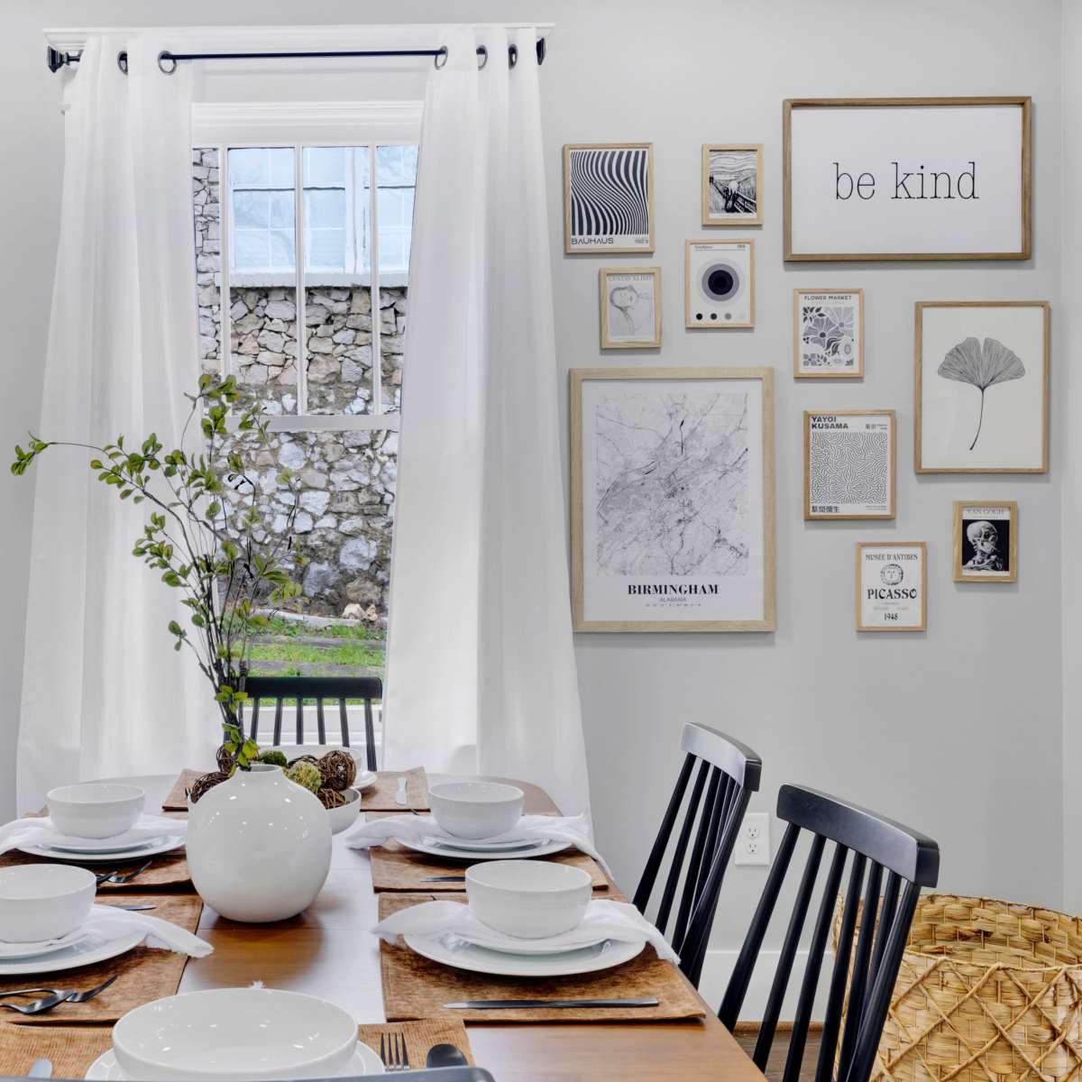 A dining room features a wooden table set for six, with white dishware and a decorative vase at its center, surrounded by wall art and a window with sheer curtains.