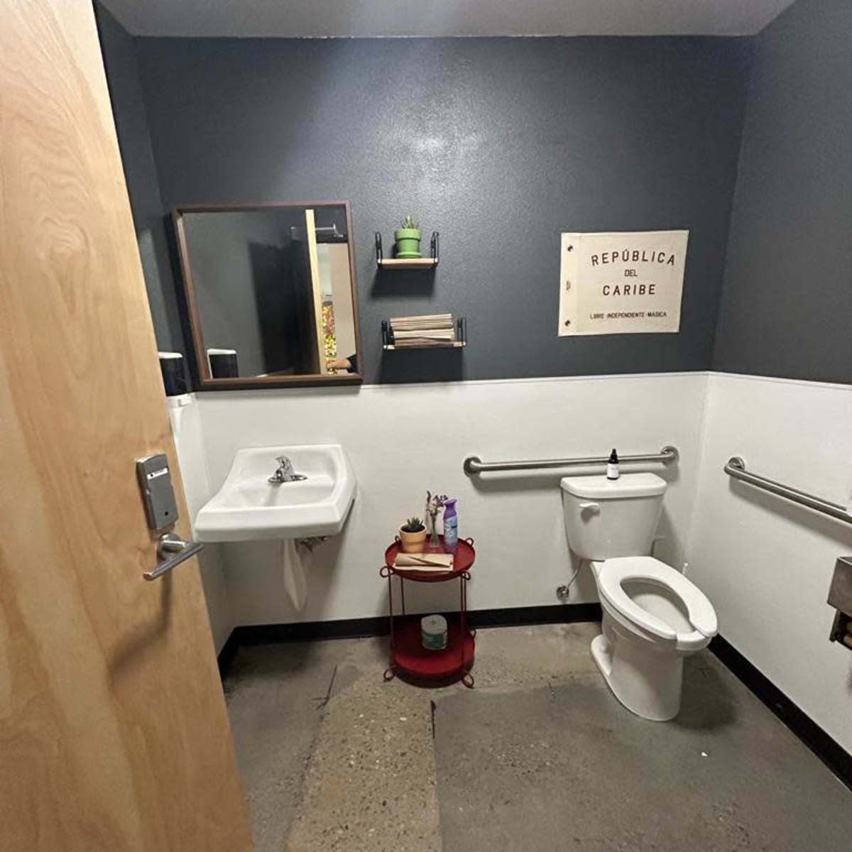 A small restroom with a sink, toilet, and a red cart holding various items, featuring gray walls and a wooden door.