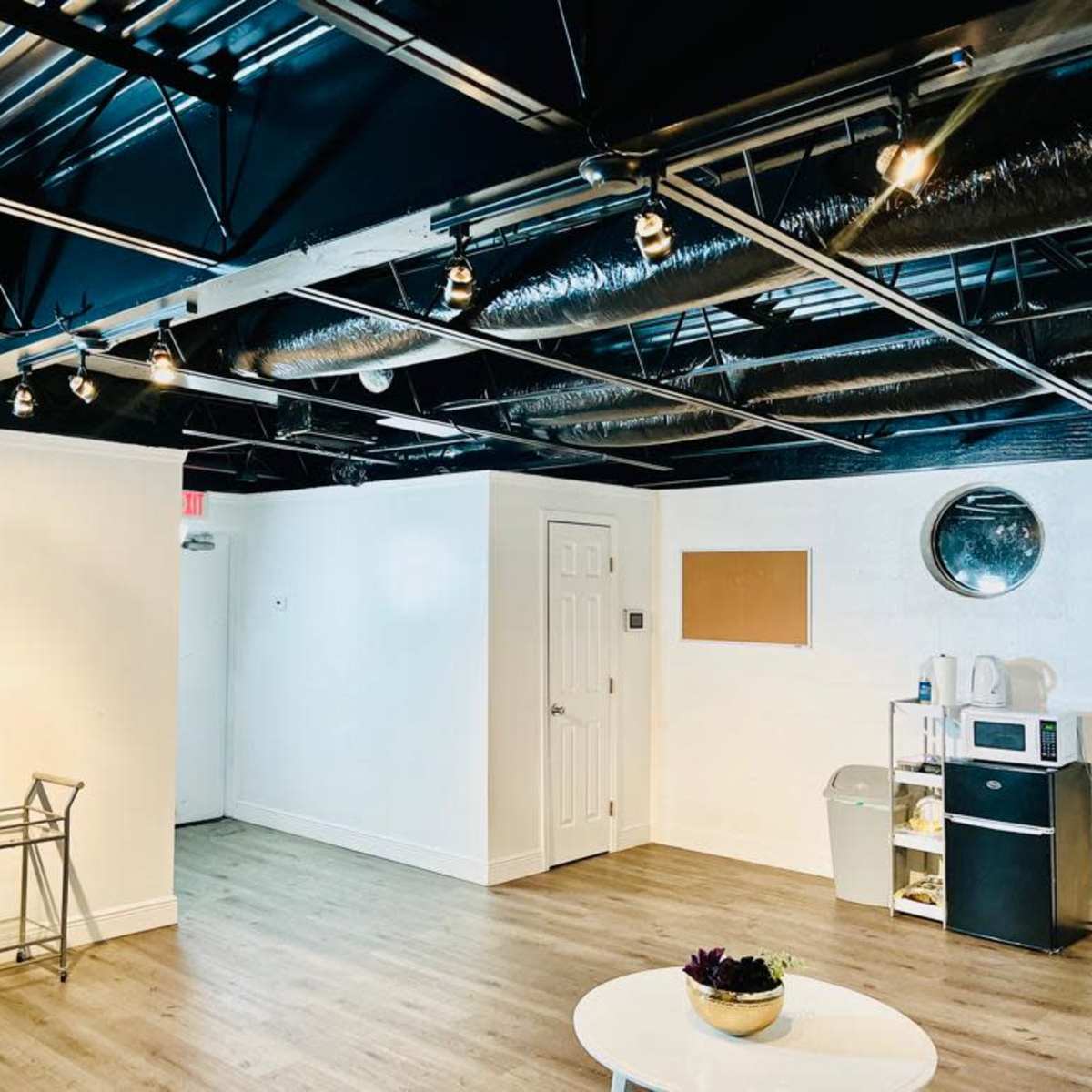 An empty room with a wooden floor, a metal frame, a door, a round mirror, a bulletin board, and kitchen appliances against one wall.