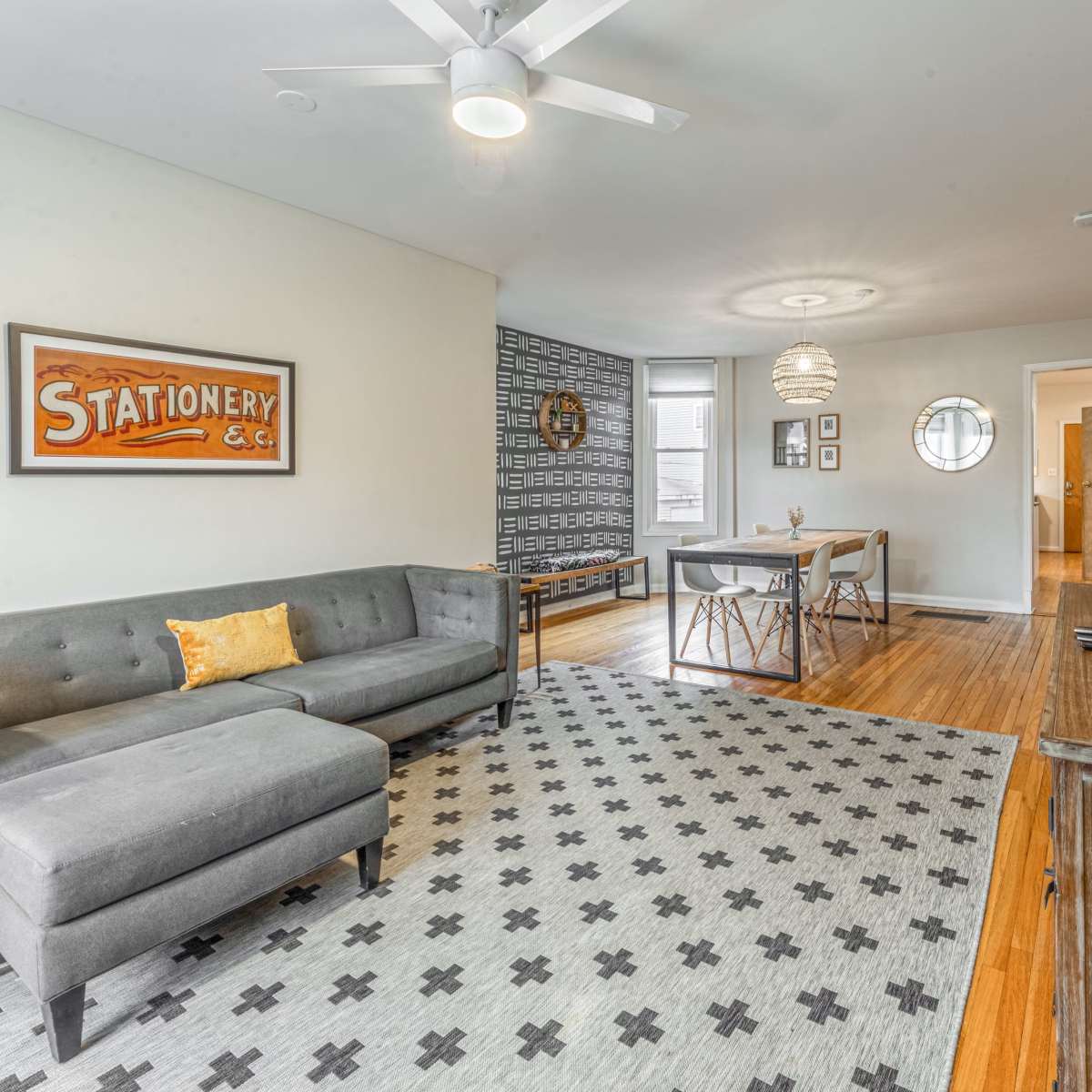 A living room with a gray sectional sofa, a patterned area rug, a dining area with a table and chairs, and a wall-mounted television.