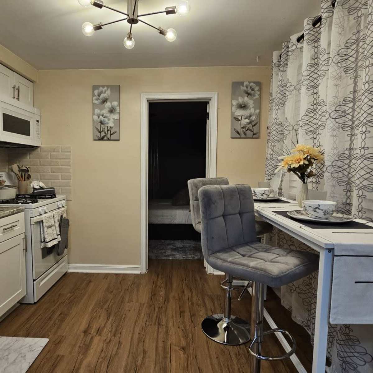 A modern kitchen with white cabinetry, a grey countertop, a small dining area for two, and a doorway leading to a darker room.
