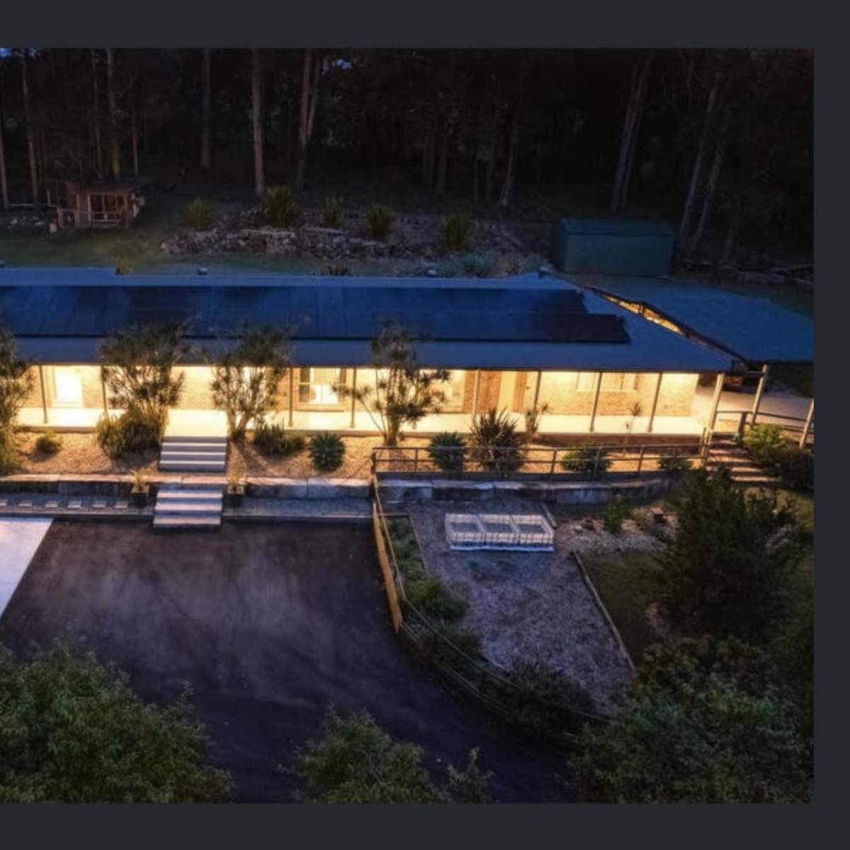A modern building with illuminated windows sits amidst trees in a dark setting, featuring a driveway and landscaped area in the foreground.