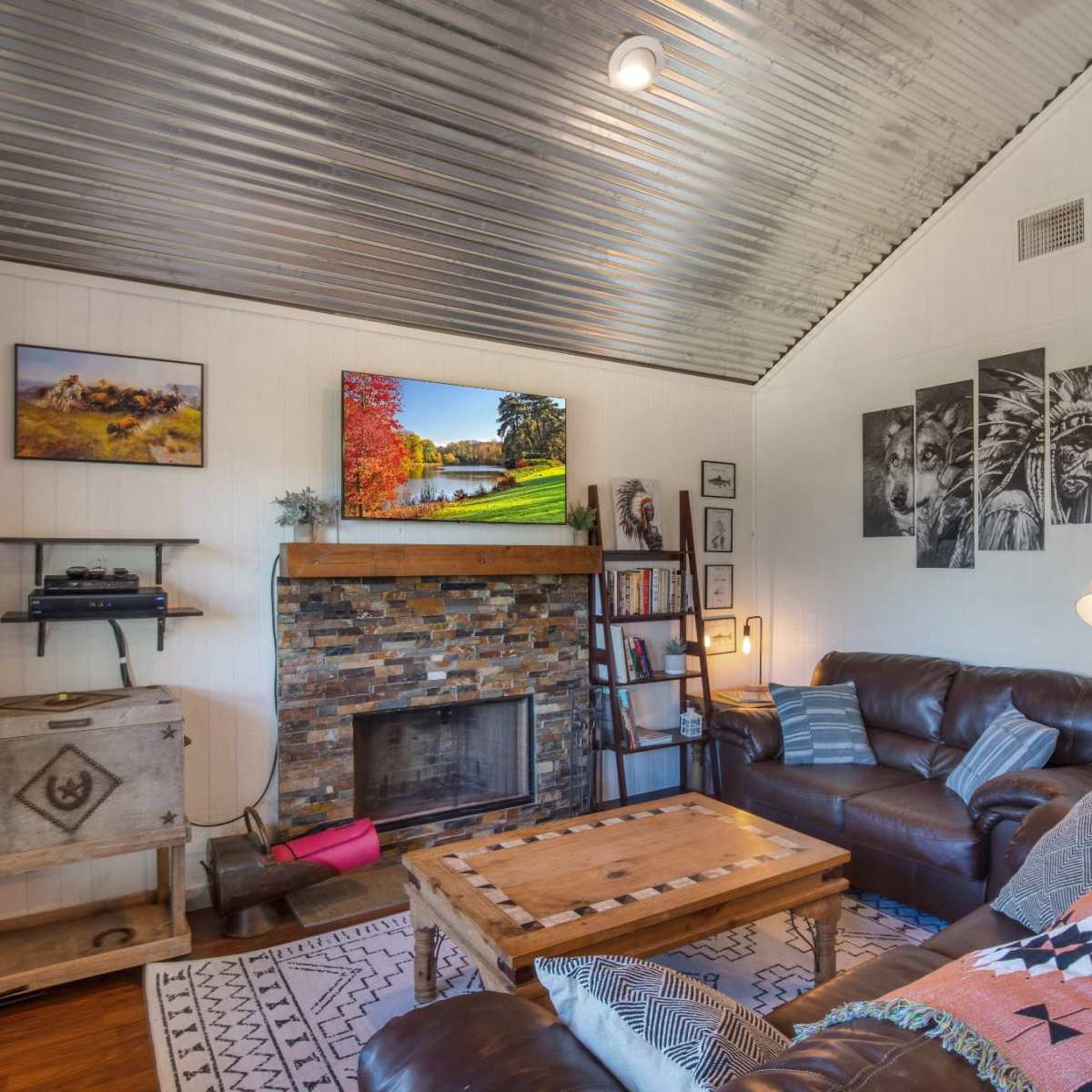 A cozy living room with a brick fireplace, a wooden coffee table, leather sofas, and artwork on the walls, featuring a television mounted above the fireplace.