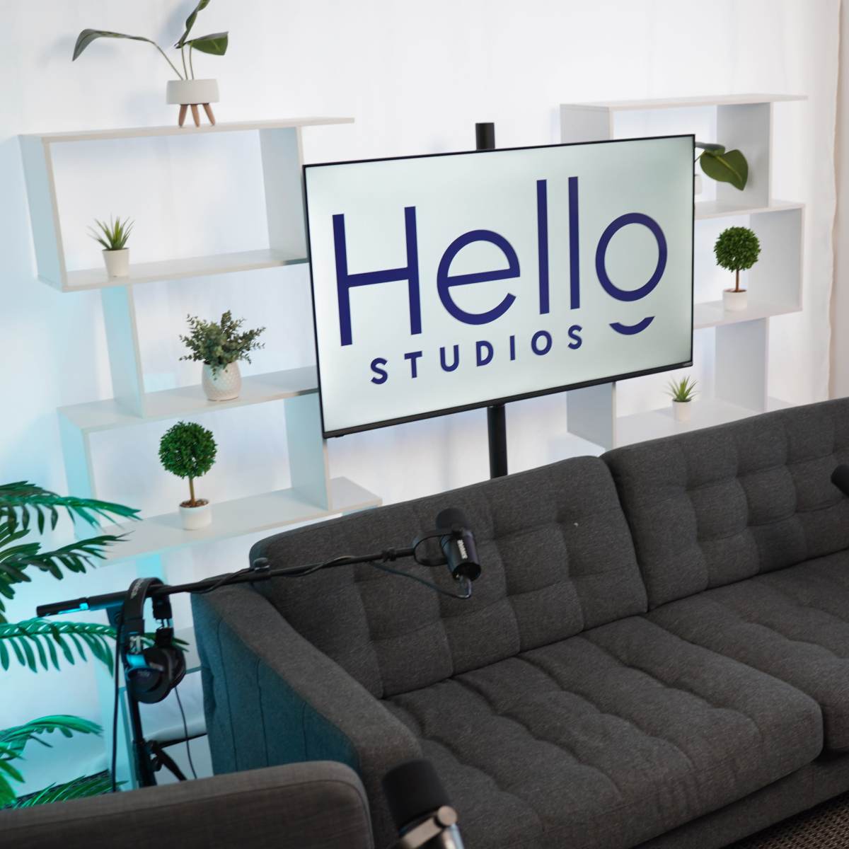 A modern studio setup featuring a gray sofa, microphones, and a large screen displaying "Hello Studios," with plants and shelves in the background.
