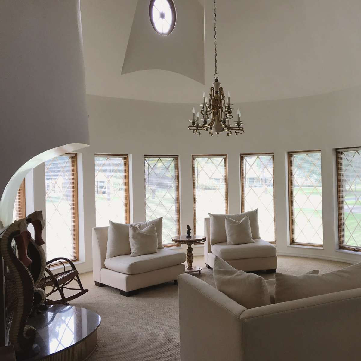 A circular living room features a chandelier, two armchairs, and a large sectional couch arranged around a coffee table, with tall windows providing natural light.