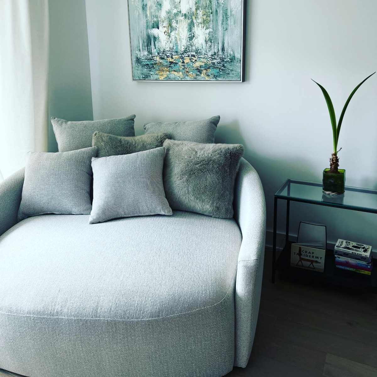 A comfortable, rounded sofa is adorned with several gray pillows, positioned next to a glass side table and a large piece of abstract artwork on the wall.