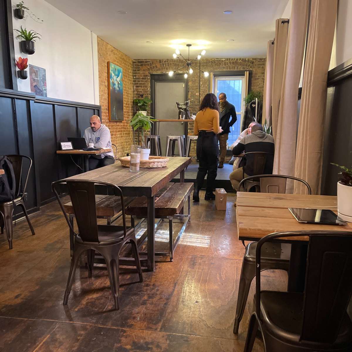 A café interior features wooden tables and metal chairs, with several patrons engaged in different activities, including one working on a laptop.