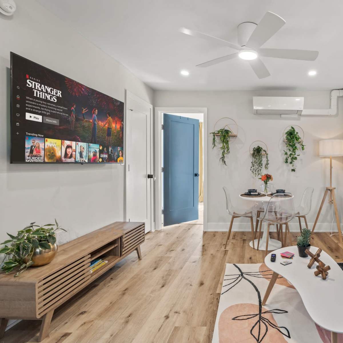 A modern living room with a light gray sofa, a coffee table, a wooden TV unit displaying a "Stranger Things" poster, and decor featuring plants against a white wall.