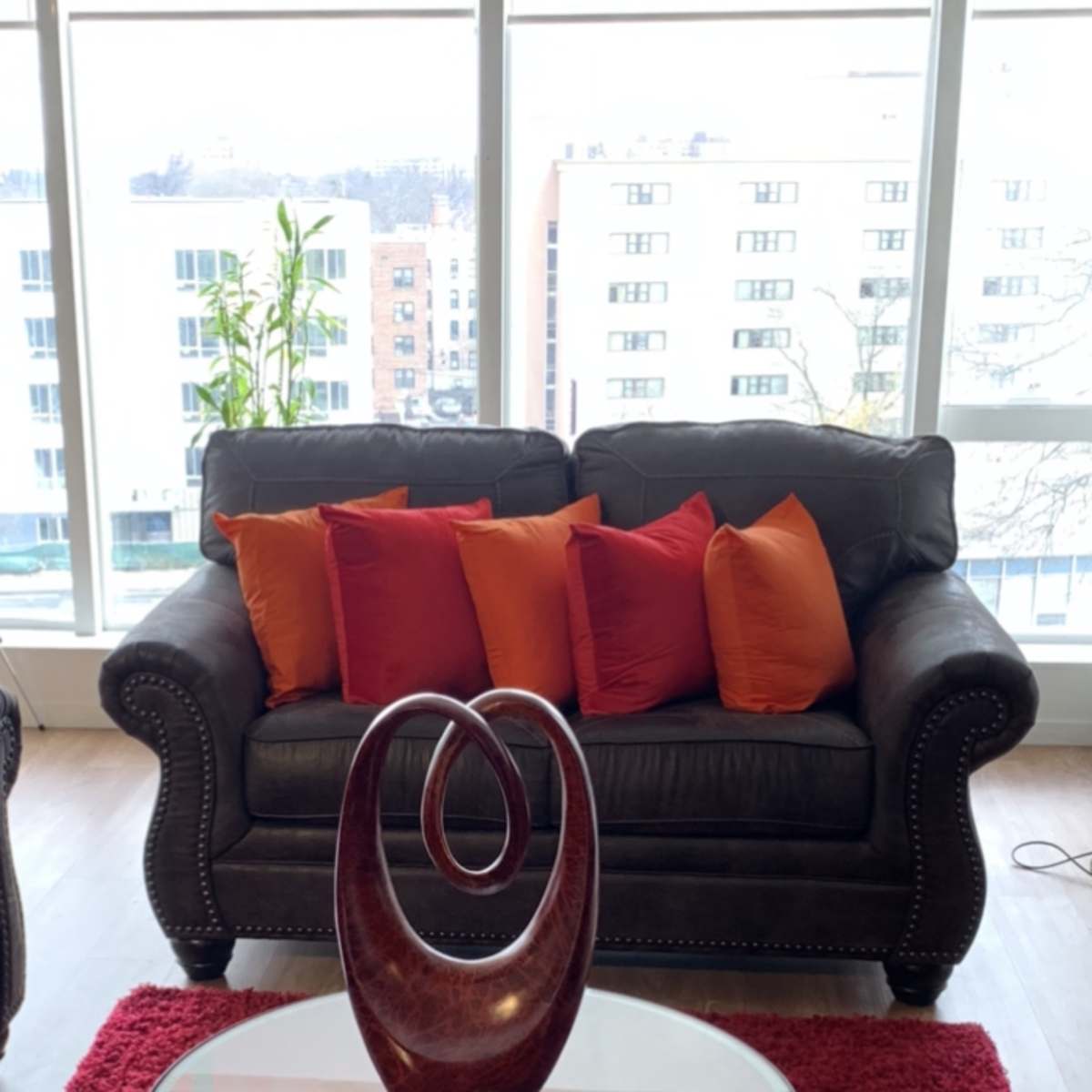 A leather couch with orange and red pillows sits in front of a glass coffee table on a red rug, with a decorative wooden sculpture and a candle on the table.
