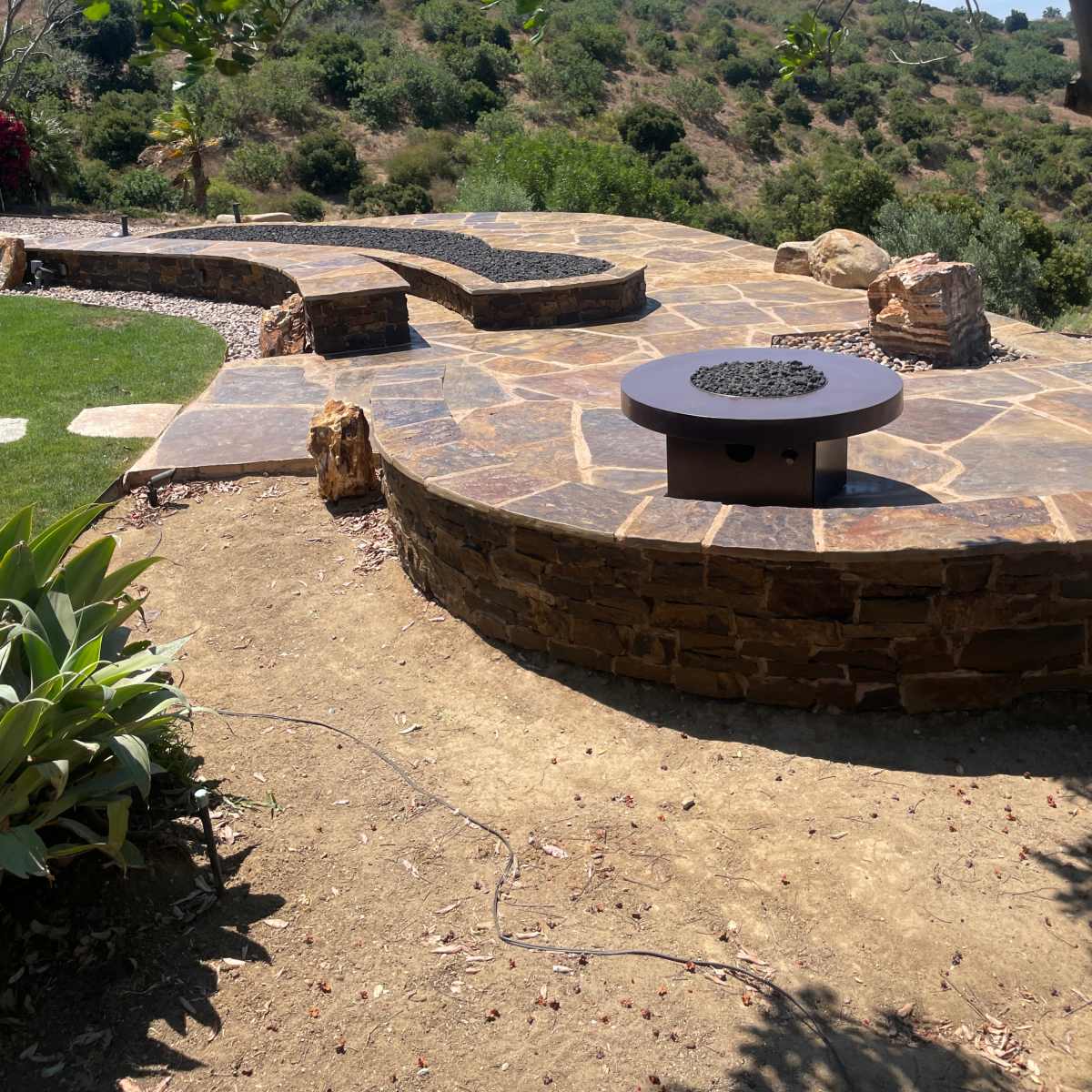 A landscaped outdoor area with a stone seating feature, surrounded by greenery and a fire pit at the center.